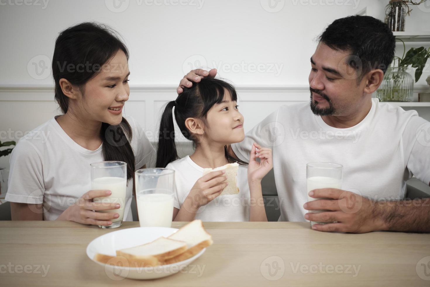 een gezond Aziatisch Thais familie, een weinig dochter, en jong ouders drinken vers wit melk in glas en brood vreugde samen Bij een dining tafel in ochtend, welzijn voeding huis ontbijt maaltijd levensstijl. foto