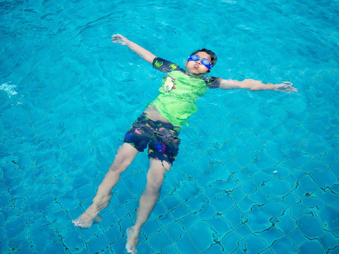 jongen vervelend een zwempak en bril zwemmen in de midden- van de zwembad met een blauw water achtergrond foto