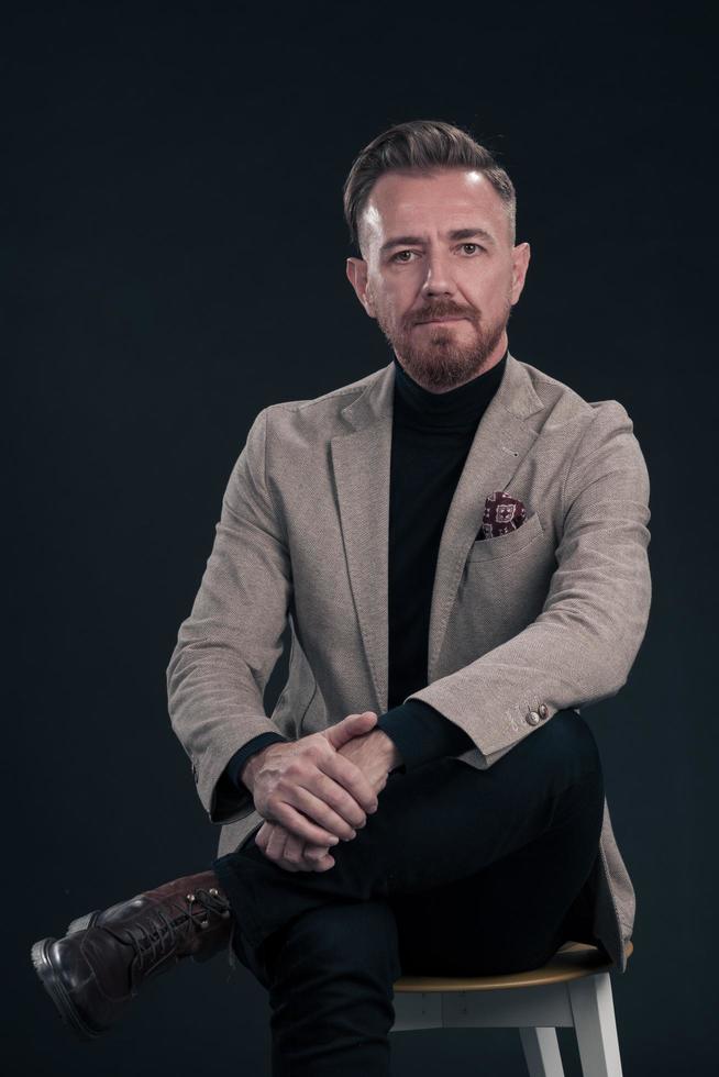 portret van volwassen zakenman vervelend modieus pak en zittend in modern studio Aan elegant stoel tegen de zwart achtergrond foto