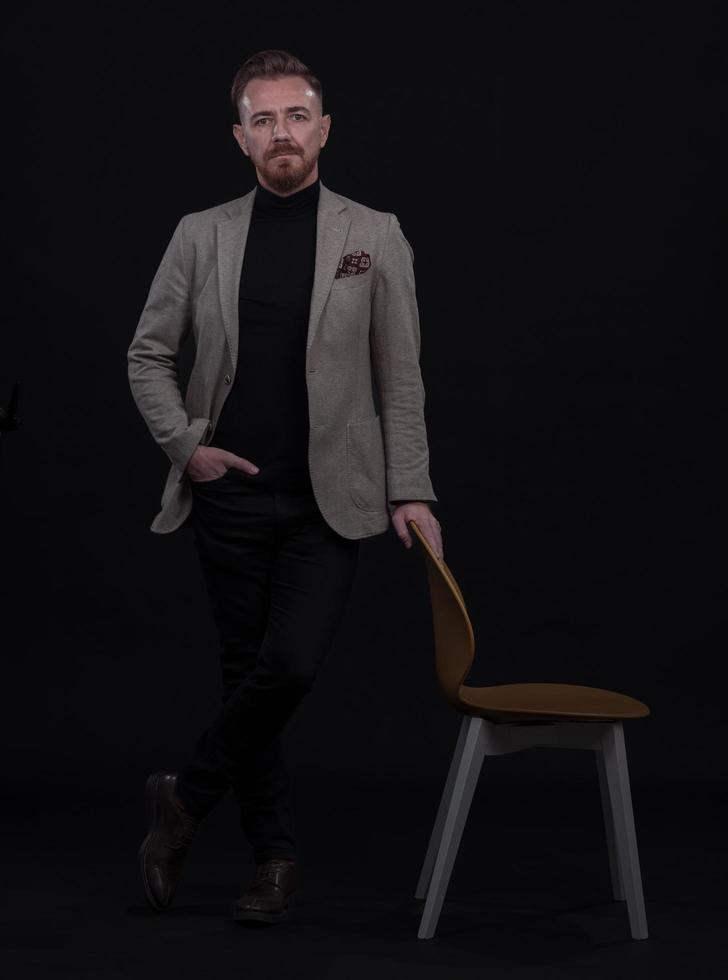 portret van volwassen zakenman vervelend modieus pak en zittend in modern studio Aan elegant stoel tegen de zwart achtergrond foto