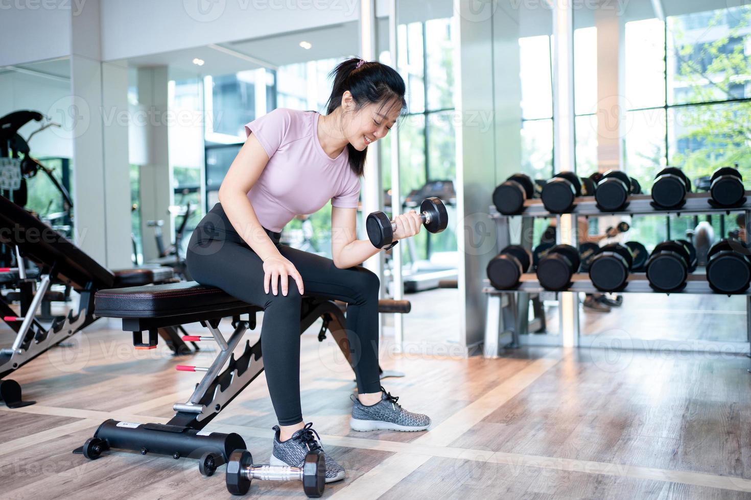 mooi atletisch gespierd Aziatisch vrouw pompen omhoog de spieren door een arm liften halter oefening Aan bank in geschiktheid Sportschool. foto