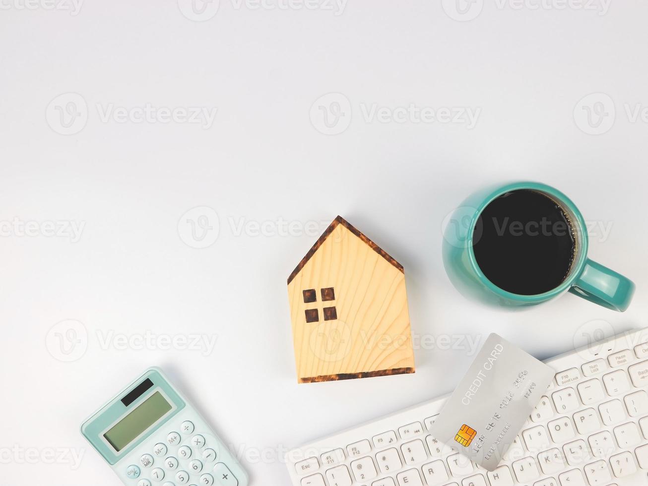 vlak lay-out van houten huis model- , blauw kop van zwart koffie, blauw rekenmachine, credit kaart en computer toetsenbord Aan wit achtergrond met kopiëren ruimte. huis aankoop concept. foto
