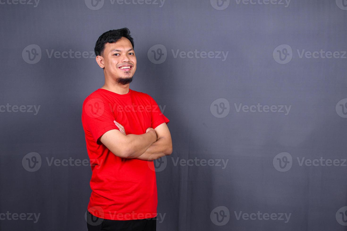 portret van aantrekkelijk Aziatisch Mens in rood t-shirt staand met gekruiste armen geïsoleerd Aan grijs achtergrond foto