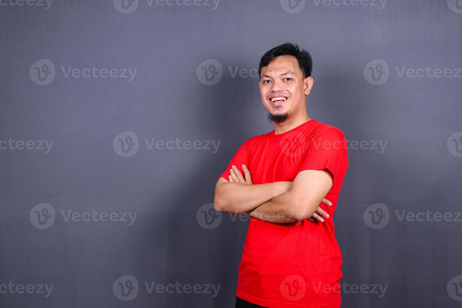 portret van aantrekkelijk Aziatisch Mens in rood t-shirt staand met gekruiste armen geïsoleerd Aan grijs achtergrond foto