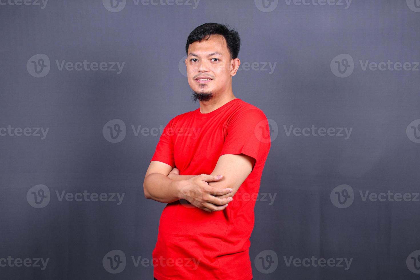 portret van aantrekkelijk Aziatisch Mens in rood t-shirt staand met gekruiste armen geïsoleerd Aan grijs achtergrond foto