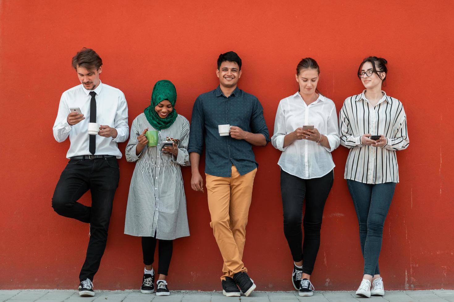 multi-etnisch groep van gewoontjes mensen uit het bedrijfsleven gebruik makend van smartphone gedurende een koffie breken van werk in voorkant van de rood muur buiten. foto