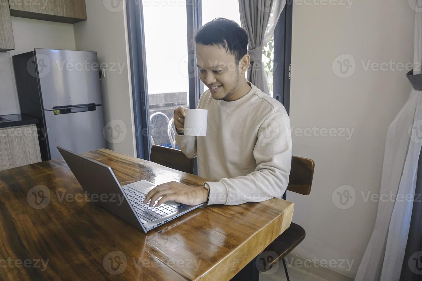 een jong Aziatisch Mens besteedt zijn tijd Bij huis zittend in de dining kamer gelukkig werken Aan zijn laptop terwijl Holding een mok foto