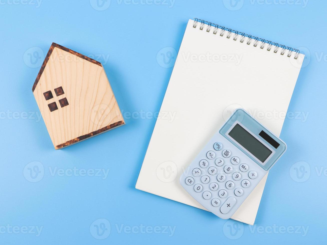 vlak lay-out van houten huis model- , geopend blanco bladzijde notitieboekje en blauw rekenmachine Aan blauw achtergrond met kopiëren ruimte. huis aankoop concept. foto
