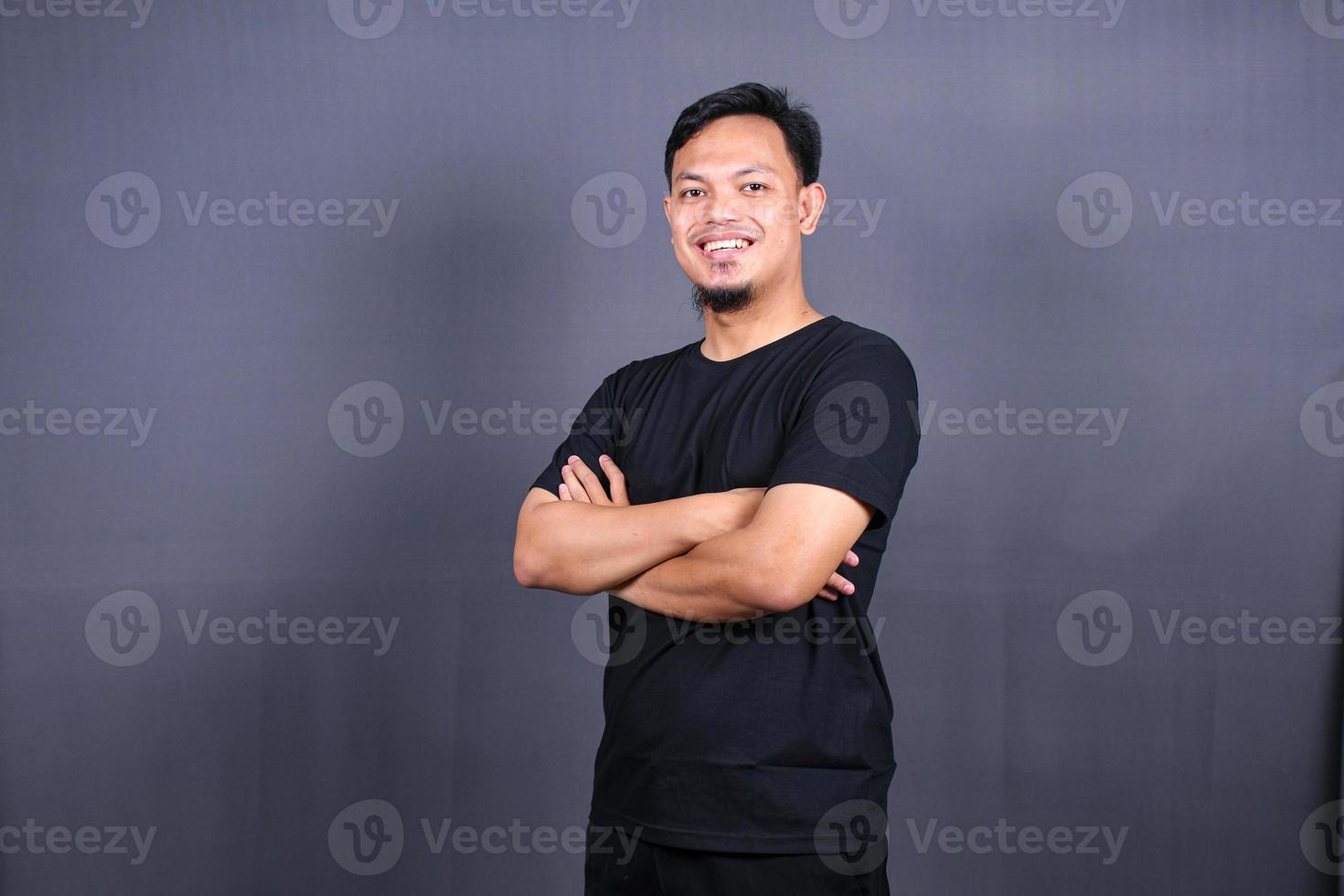 glimlachen knap Aziatisch Mens in zwart t-shirt staand met gekruiste armen geïsoleerd Aan grijs achtergrond foto