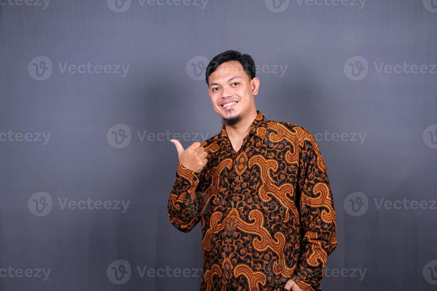 portret van een gelukkig jong Mens in batik Indonesië richten vingers weg Bij kopiëren ruimte geïsoleerd over- grijs achtergrond foto