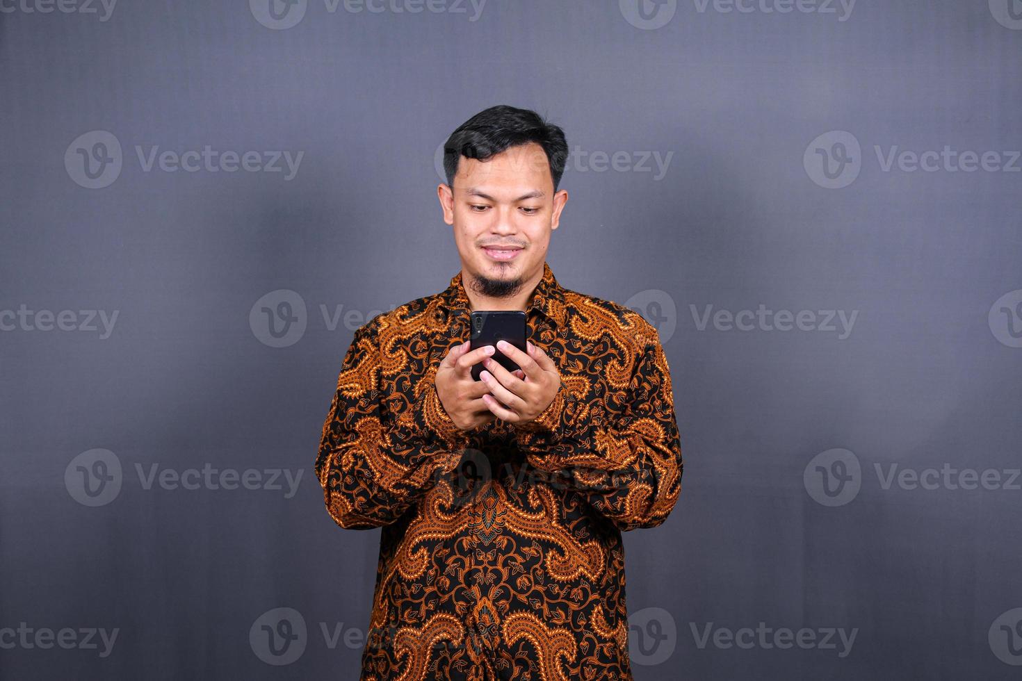 portret van Aziatisch jong Mens vervelend batik gebruik makend van mobiele telefoon Aan grijs achtergrond foto