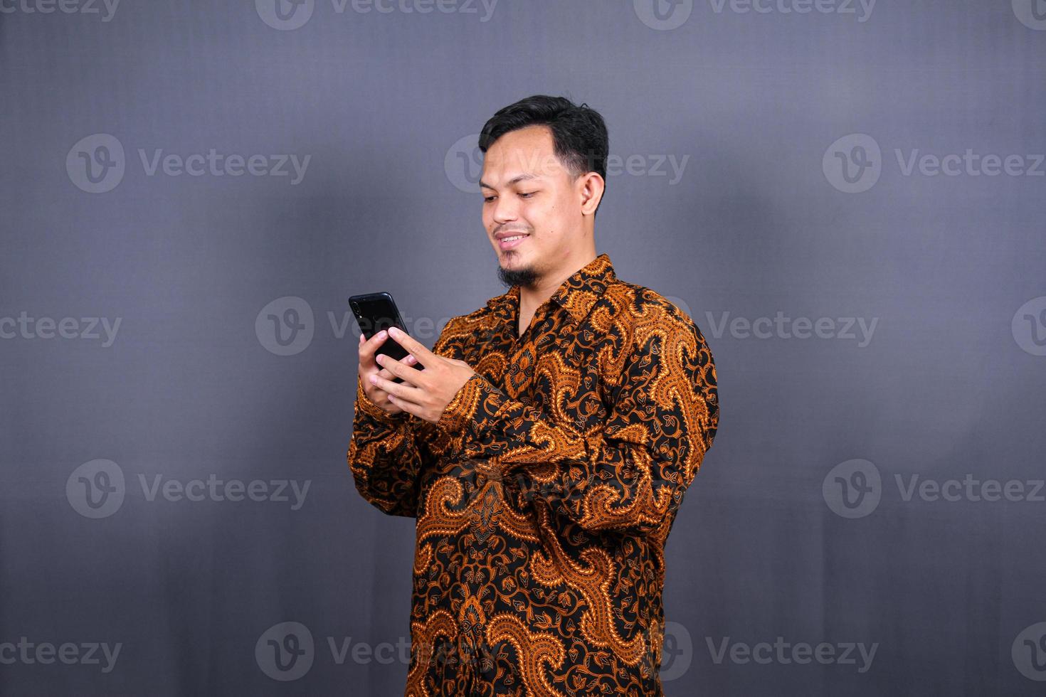 portret van Aziatisch jong Mens vervelend batik gebruik makend van mobiele telefoon Aan grijs achtergrond foto