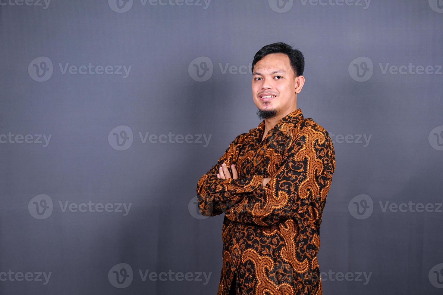 portret van aantrekkelijk jong Aziatisch Mens in batik Indonesië staand met gekruiste armen geïsoleerd Aan grijs achtergrond foto