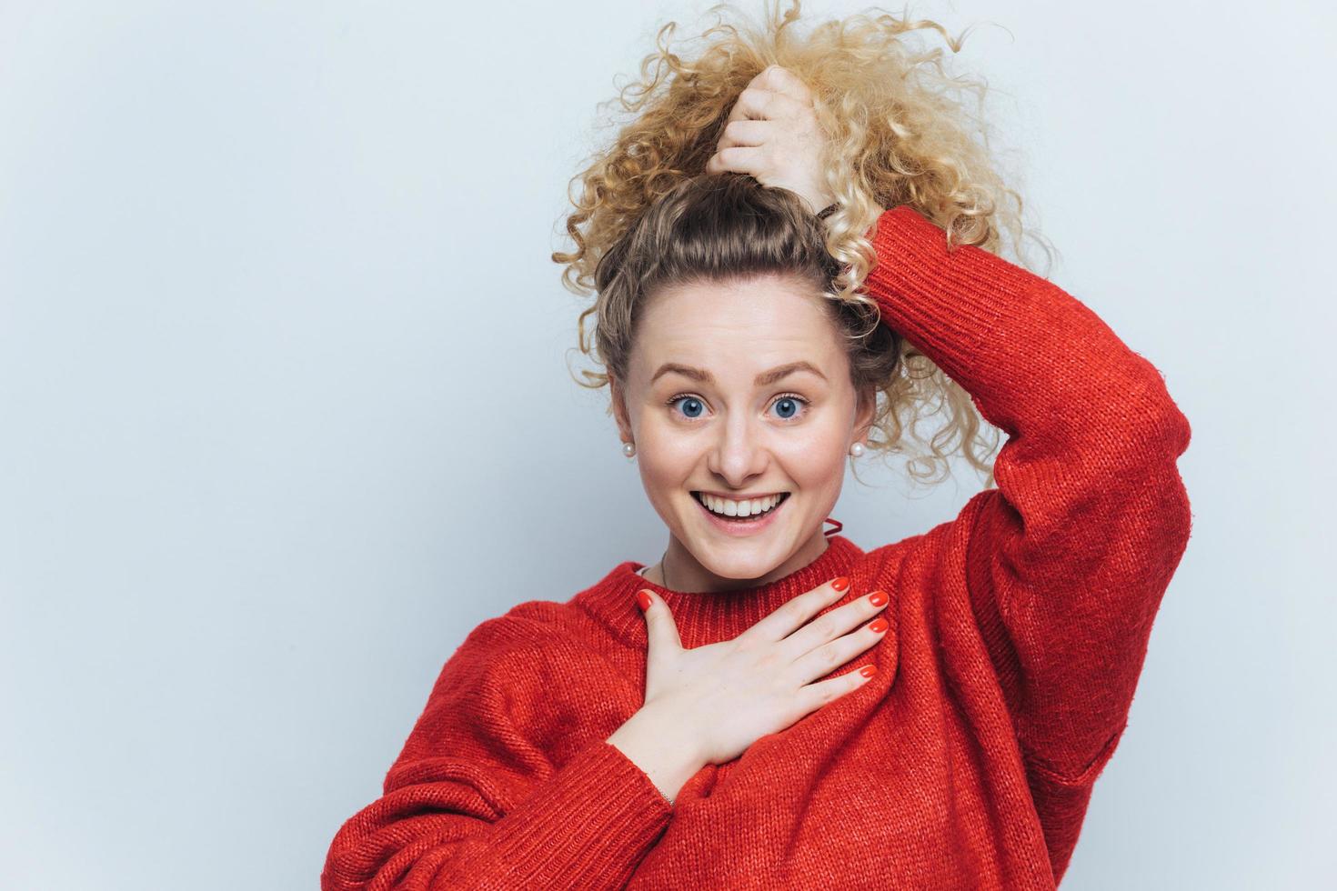 schattige opgewonden jonge vrouw met krullend haar, houdt haar vastgebonden in paardenstaart, gekleed in casual rode trui, blij om positief nieuws van gesprekspartner te ontvangen, poseert tegen witte studio-achtergrond. foto