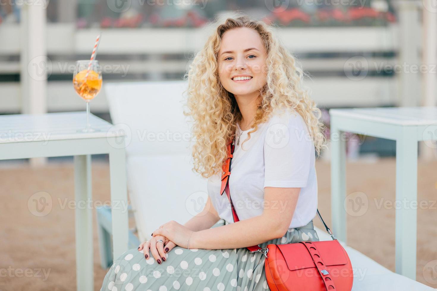 mensen en recreatie concept. mooie jonge vrouw met krullend borstelig haar, draagt een t-shirt en rok, heeft een lichte kleine tas, rust buiten, drinkt een verse cocktail, kijkt positief direct naar de camera foto