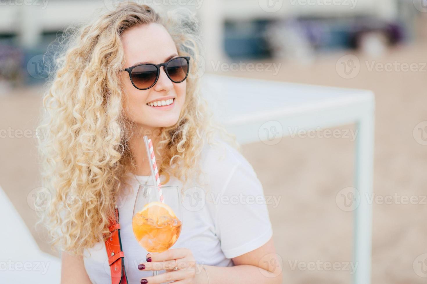 positieve blonde vrouw met krullend haar, draagt een zonnebril, kijkt met een gelukkige uitdrukking opzij, heeft een zachte glimlach, houdt een verse cocktail vast, wacht op een vriend op het strand. mensen, resort, recreatieconcept foto