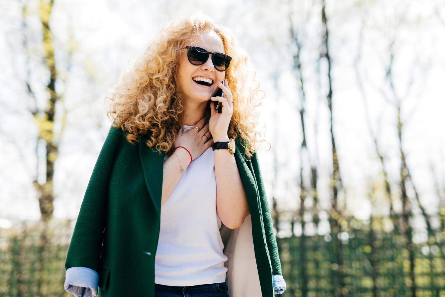 half profiel portret van aantrekkelijke vrouw met krullend haar, gekleed in stijlvolle kleding en zonnebril pratend op smartphone, glimlachend en lachend met een blije en opgewonden uitdrukking die haar nek aanraakt foto