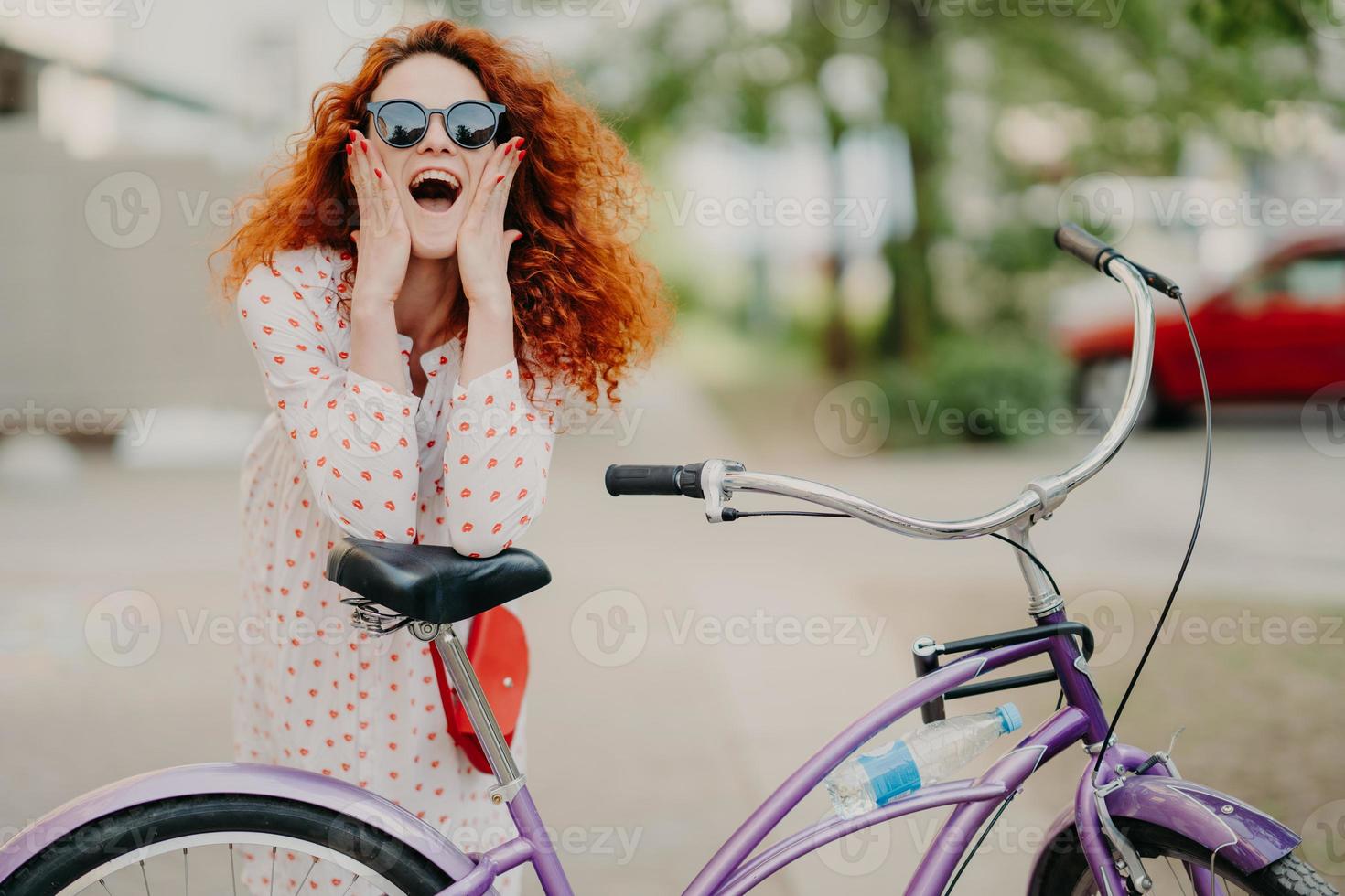 vrolijke vrouwelijke stadsfietser draagt een zonnebril en jurk, heeft luxueus fris foxyhaar, houdt de ellebogen op het zadel van de fiets, brengt recreatietijd door in de open lucht, poseert over een wazige straatachtergrond foto