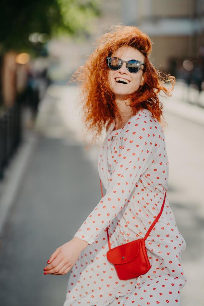 gelukkige zorgeloze roodharige vrouw lacht positief, brengt vrije tijd buiten door in de stad, draagt een zonnebril, een jurk met lange mouwen, draagt een rode tas, heeft plezier, geniet van recreatietijd. mensen en levensstijl foto