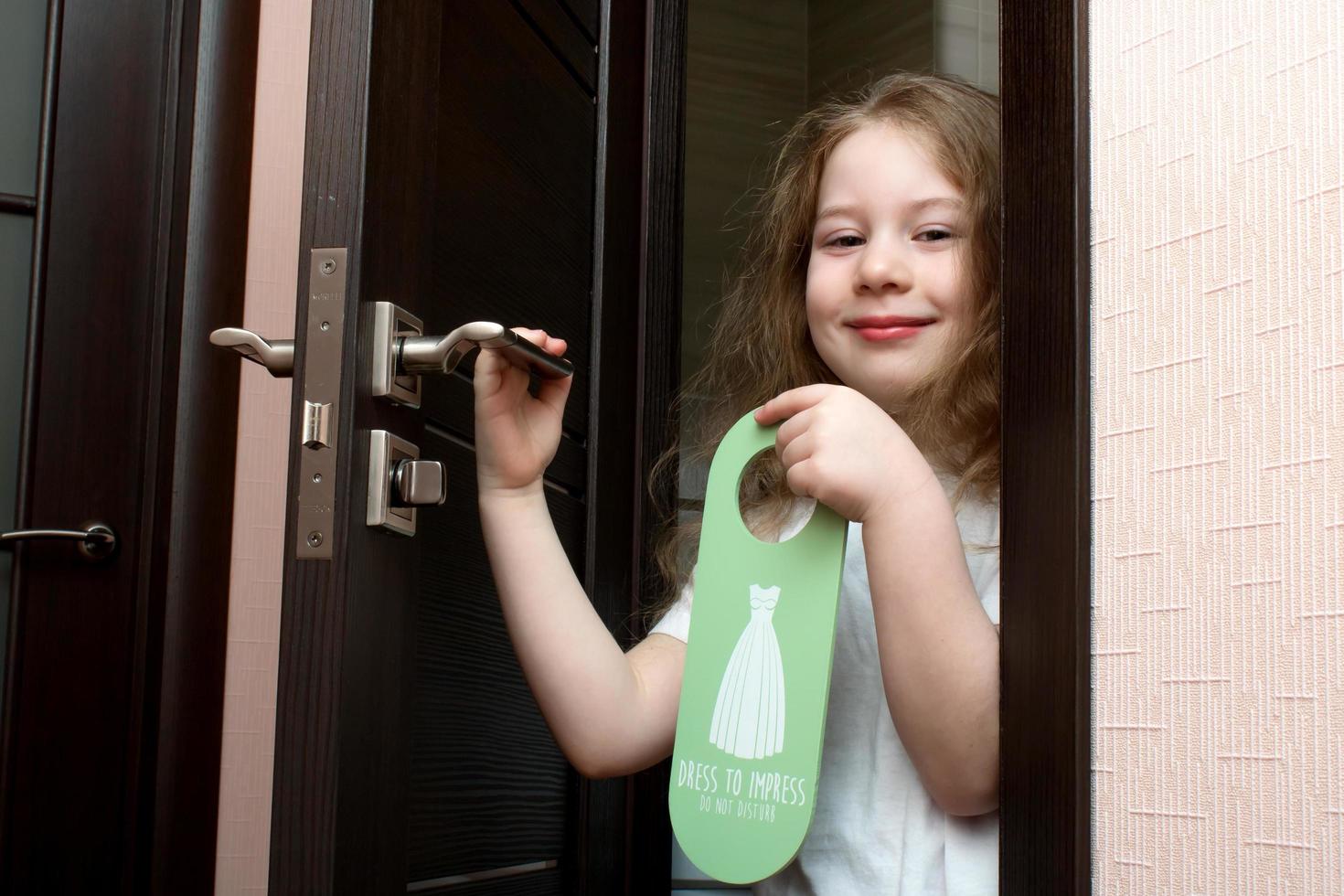 weinig meisje met geschilderd lippenstift gluurt uit van achter een deur dat zegt jurk naar indruk maken niet doen storen foto