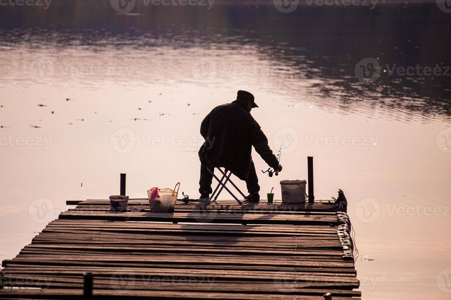 silhouet van een visser van achter zittend Aan een houten pier gedurende avond visvangst foto