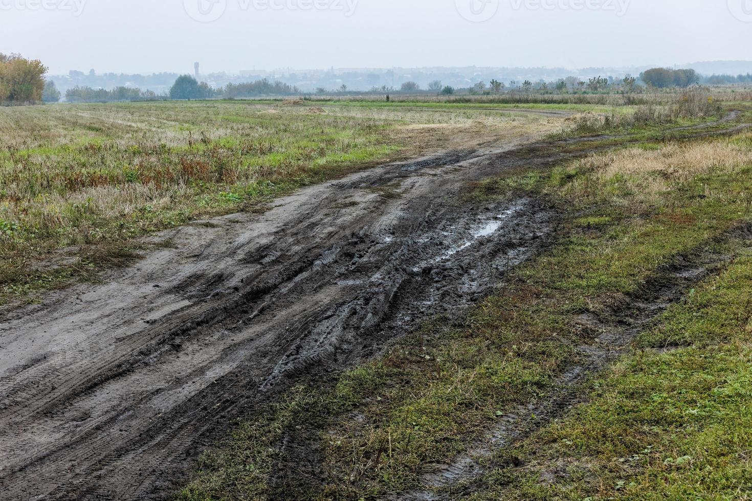 herfst- mistig ochtend- rustiek landschap met aarde weg in de voorgrond en klein gebouwen Aan heuvel in de achtergrond foto