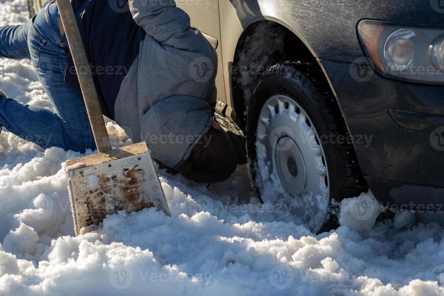 Mens werken Bij auto zit vast in sneeuw Aan knie met Schep Bij daglicht van de weg af foto