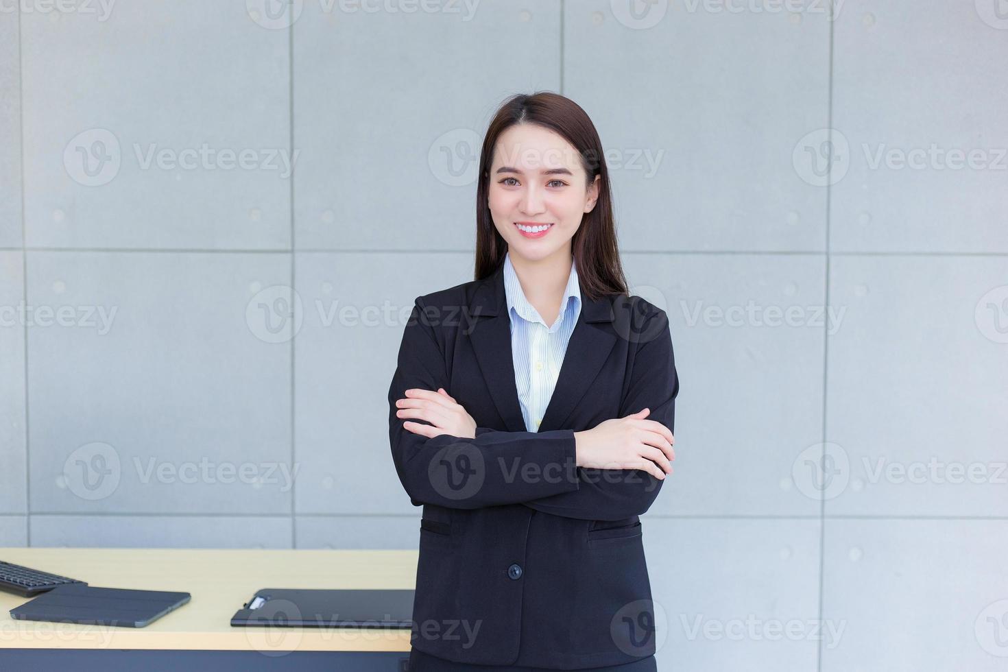 Aziatisch professioneel werken vrouw wie heeft lang haar- draagt zwart formeel pak met blauw overhemd terwijl ze arm kruispunt en zelfverzekerd glimlacht in kantoor kamer. foto