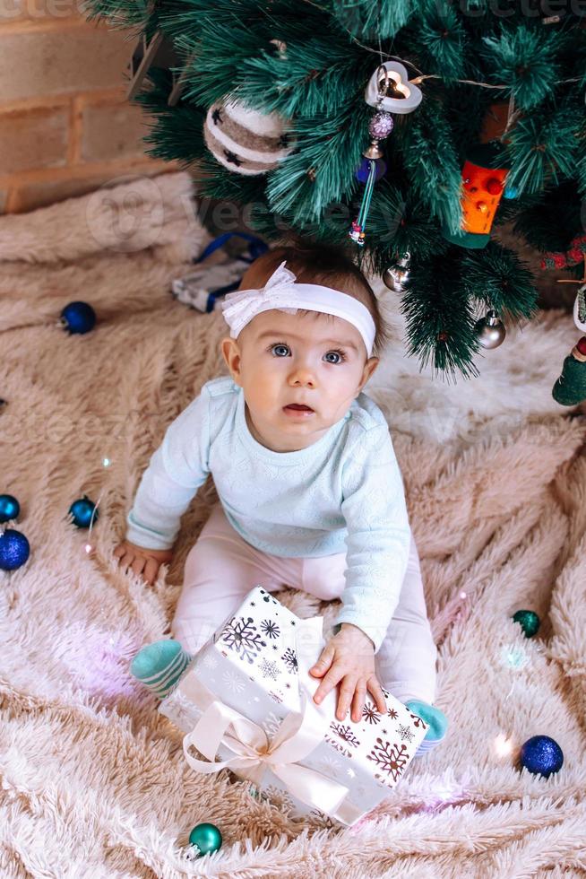 schattig weinig meisje in huis kleding van pastel kleuren is zittend in de buurt Kerstmis boom Aan een beige pluche plaid met Kerstmis decoraties en lichten en Holding in haar handen Kerstmis geschenk. foto