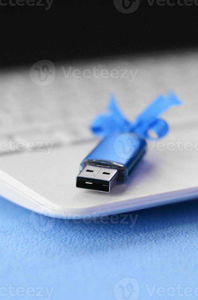 briljant blauw USB flash geheugen kaart met een blauw boog leugens Aan een deken van zacht en harig licht blauw fleece kleding stof naast naar een wit laptop. klassiek vrouw geschenk ontwerp voor een geheugen kaart foto