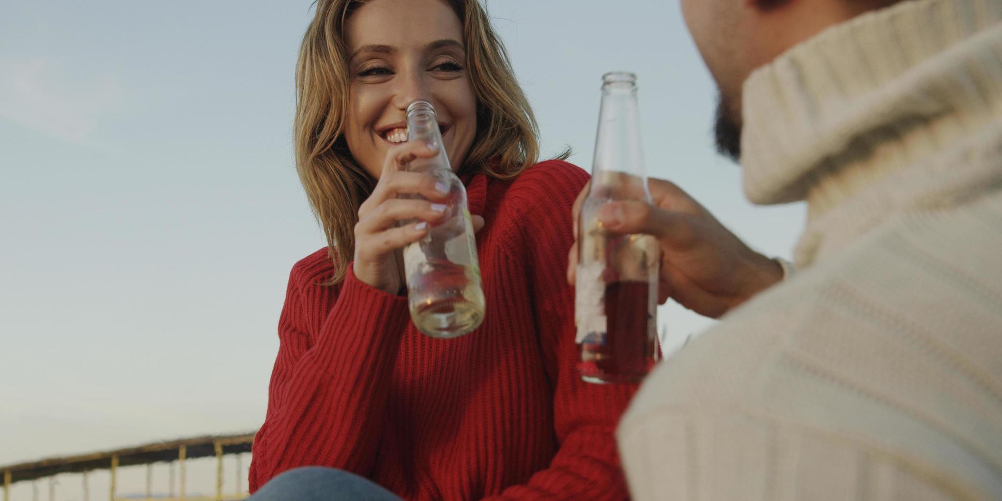 liefhebbend jong paar zittend Aan de strand naast kampvuur drinken bier foto