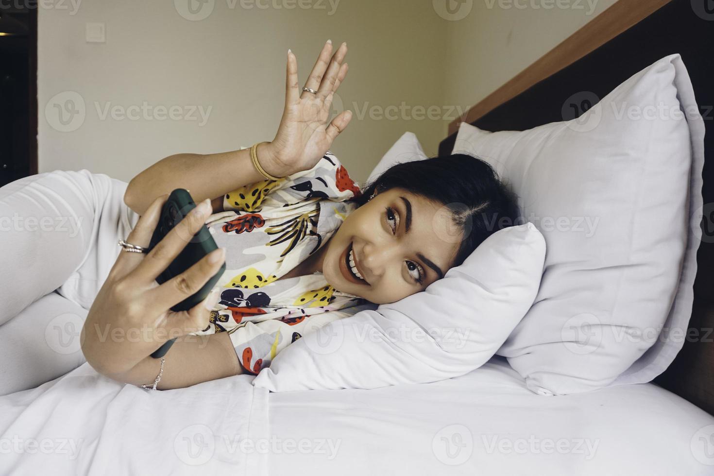 opgewonden blij jong Aziatisch vrouw aan het doen video telefoontje of selfie schot Aan mobiel telefoon golvend groet terwijl aan het liegen Aan bed foto