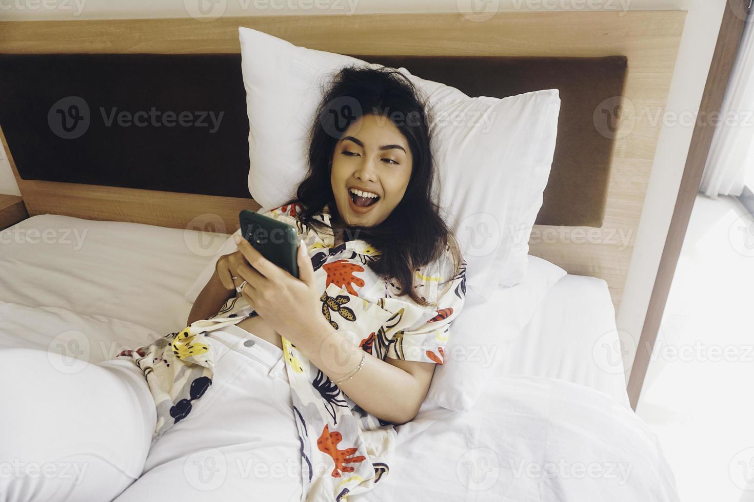 gelukkig jong vrouw controle Aan haar telefoon terwijl aan het liegen in bed foto