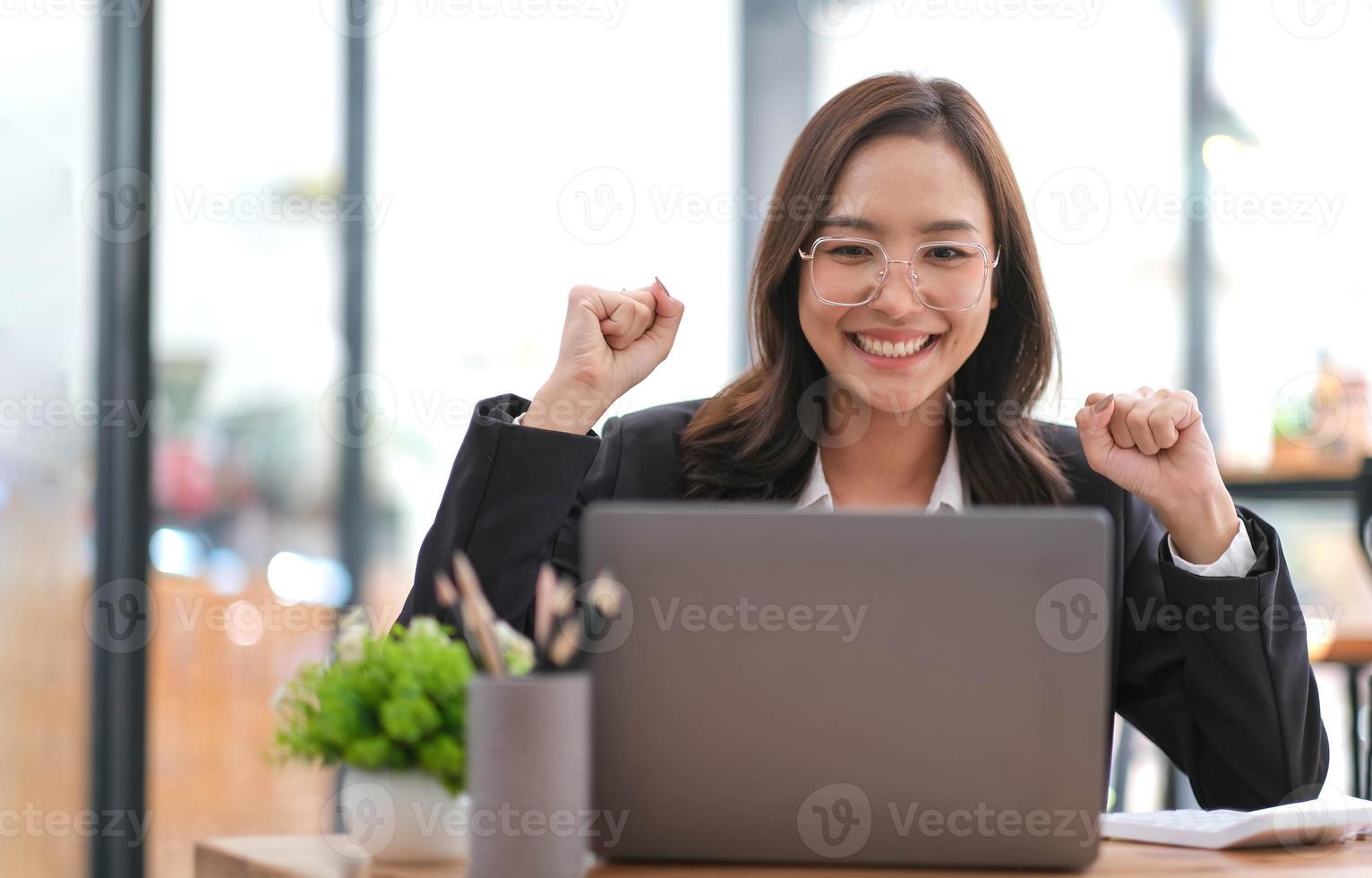 charmant en vrolijk millennial Aziatisch zakenvrouw in formeel pak zit Bij haar bureau, tonen triomf hand- gebaar, lezing een document, vieren haar succes, krijgen baan Promotie. foto