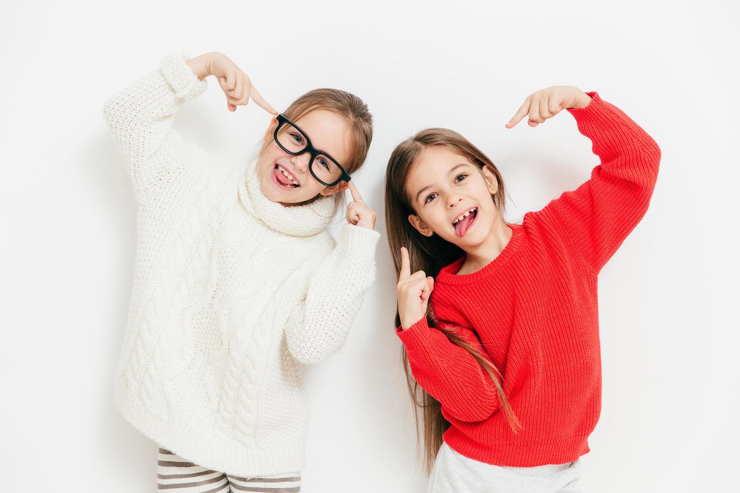 vrolijke meisjes zijn in een goed humeur, dragen een oversized trui en een bril, maken een cool teken, poseren samen tegen een witte achtergrond. gelukkige vrouwelijke kinderen hebben plezier. kindertijd en geluk concept foto