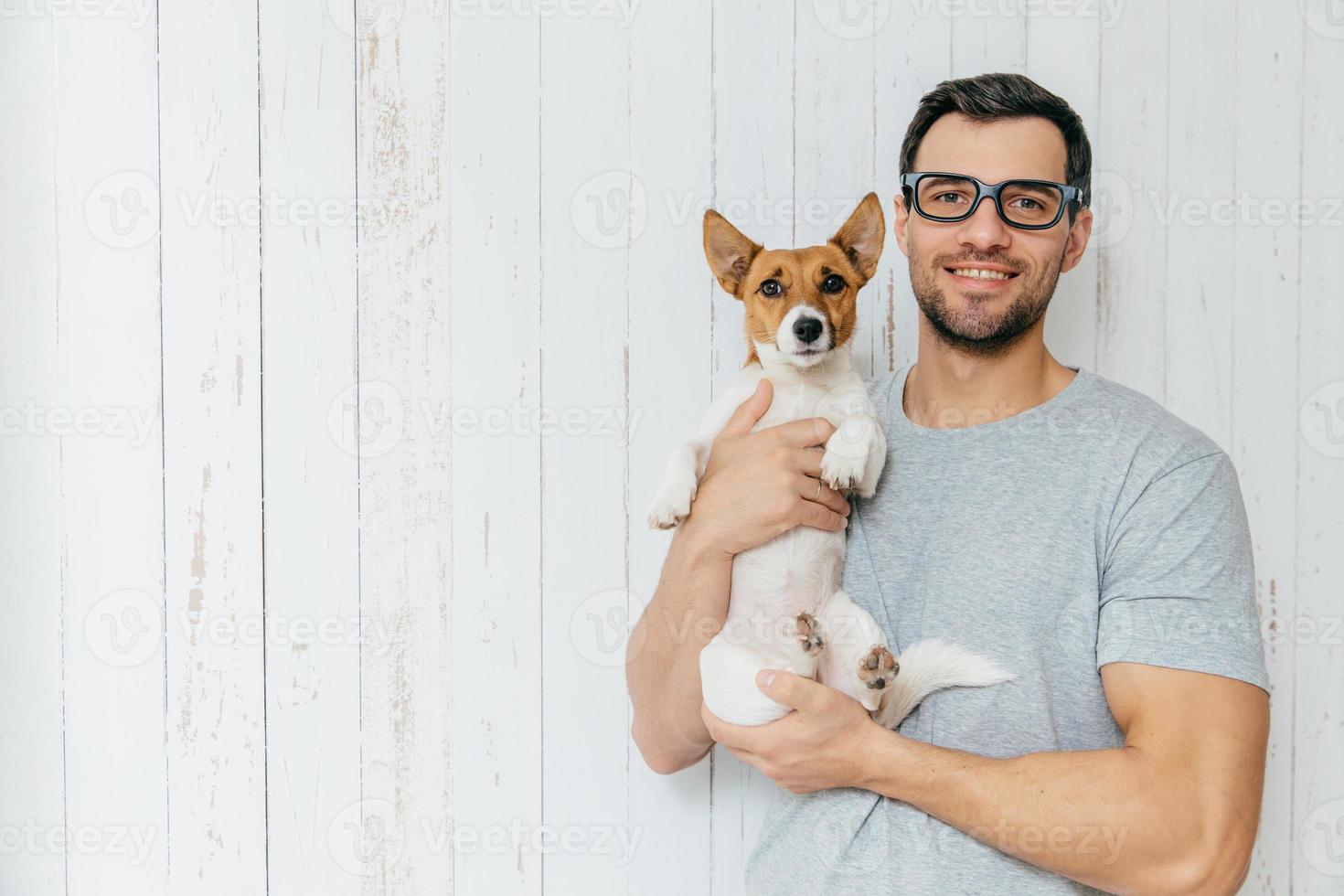 mensen, positiviteit, dieren en vriendschapsconcept. gelukkig ongeschoren mannetje draagt vrijetijdskleding, houdt puppy vast, loopt samen, staat samen tegen een witte houten muur met lege kopieerruimte foto