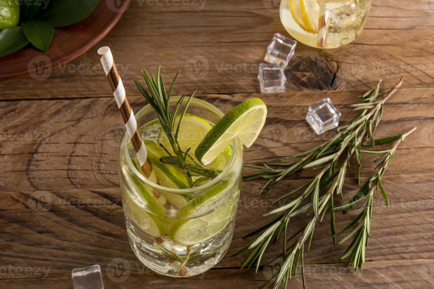 een glas met een verkoudheid verfrissend drinken met limoen, ijs kubussen en takjes van rozemarijn Aan een houten tafel. ontgiften water. foto