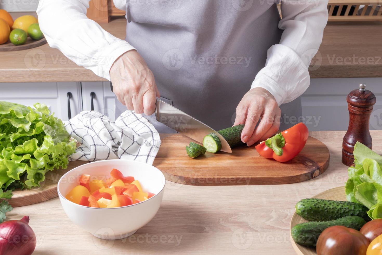 een ouderen vrouw in een grijs schort bezuinigingen een vers groen komkommer Aan een snijdend bord in een modern keuken. voorbereiding van groente salade. foto