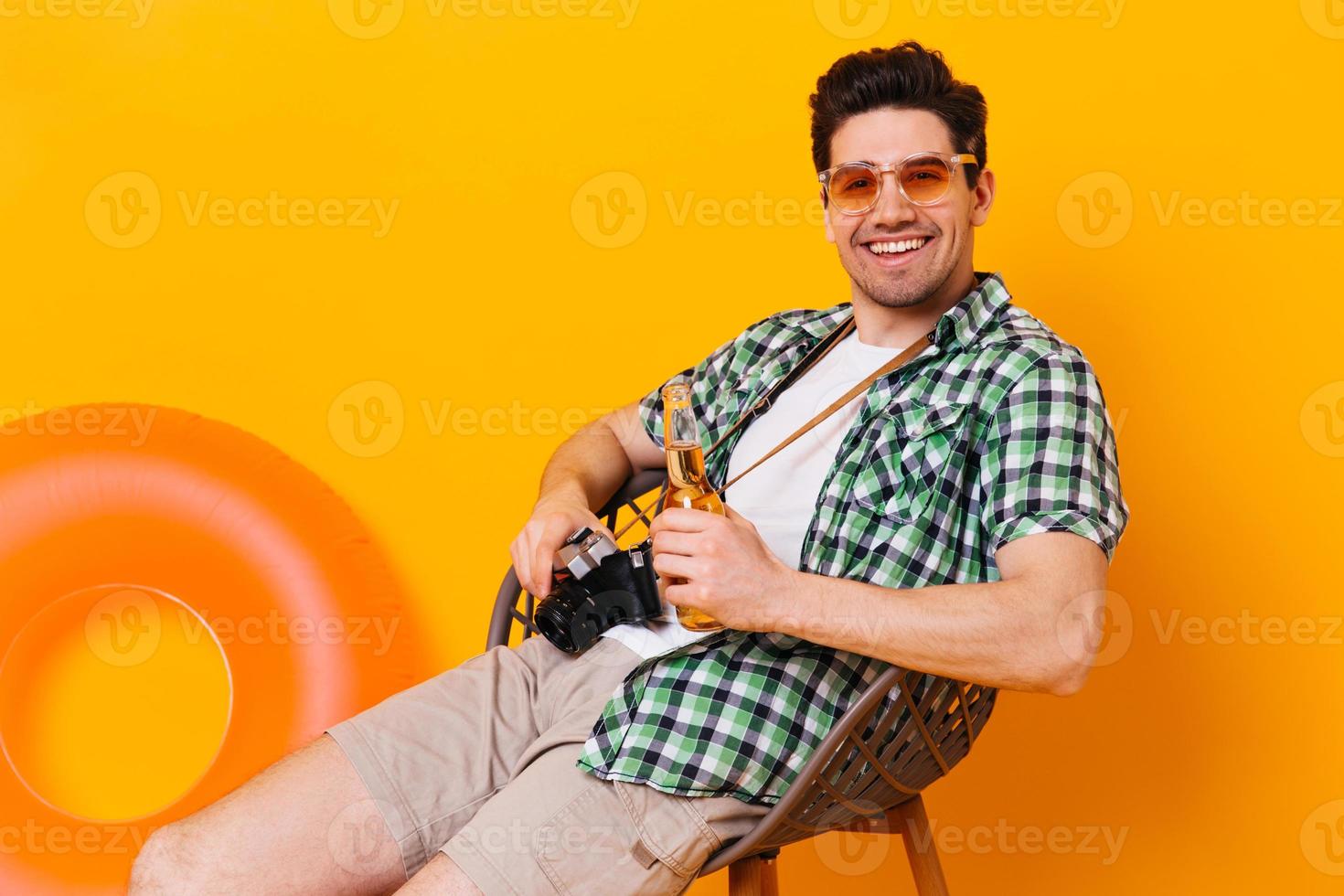gelukkig vent in oranje bril is ontspannende in houten stoel, lachend, Holding fles van bier en retro foto