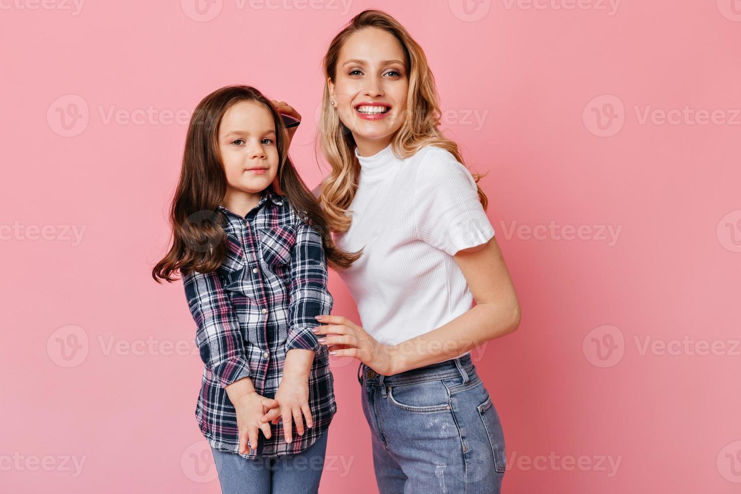 positief vrouw in wit t-shirt en jeans poetsen golvend donker haar- van dochter en poseren Aan roze ba foto