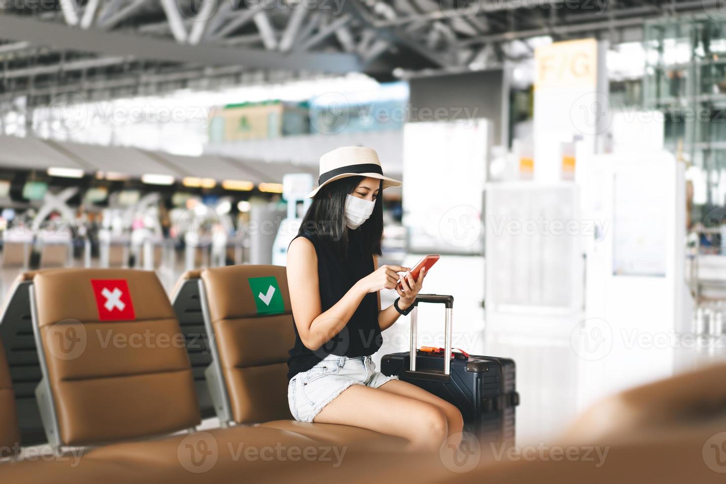 volwassen toerist vrouw met bagage slijtage masker voor covid-19 controleren in met mobiel telefoon Bij luchthaven terminal. foto