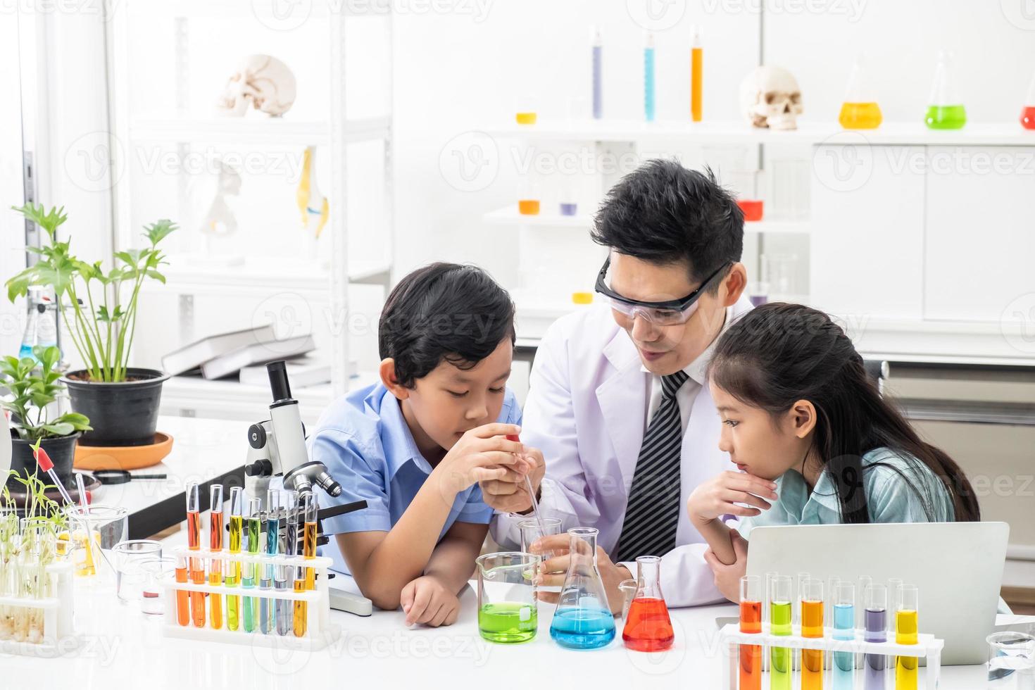 jong Aziatisch jongen en meisje glimlach en hebben pret terwijl aan het doen wetenschap experiment in laboratorium klas met docent. studie met wetenschappelijk uitrusting en buizen. onderwijs concept. foto