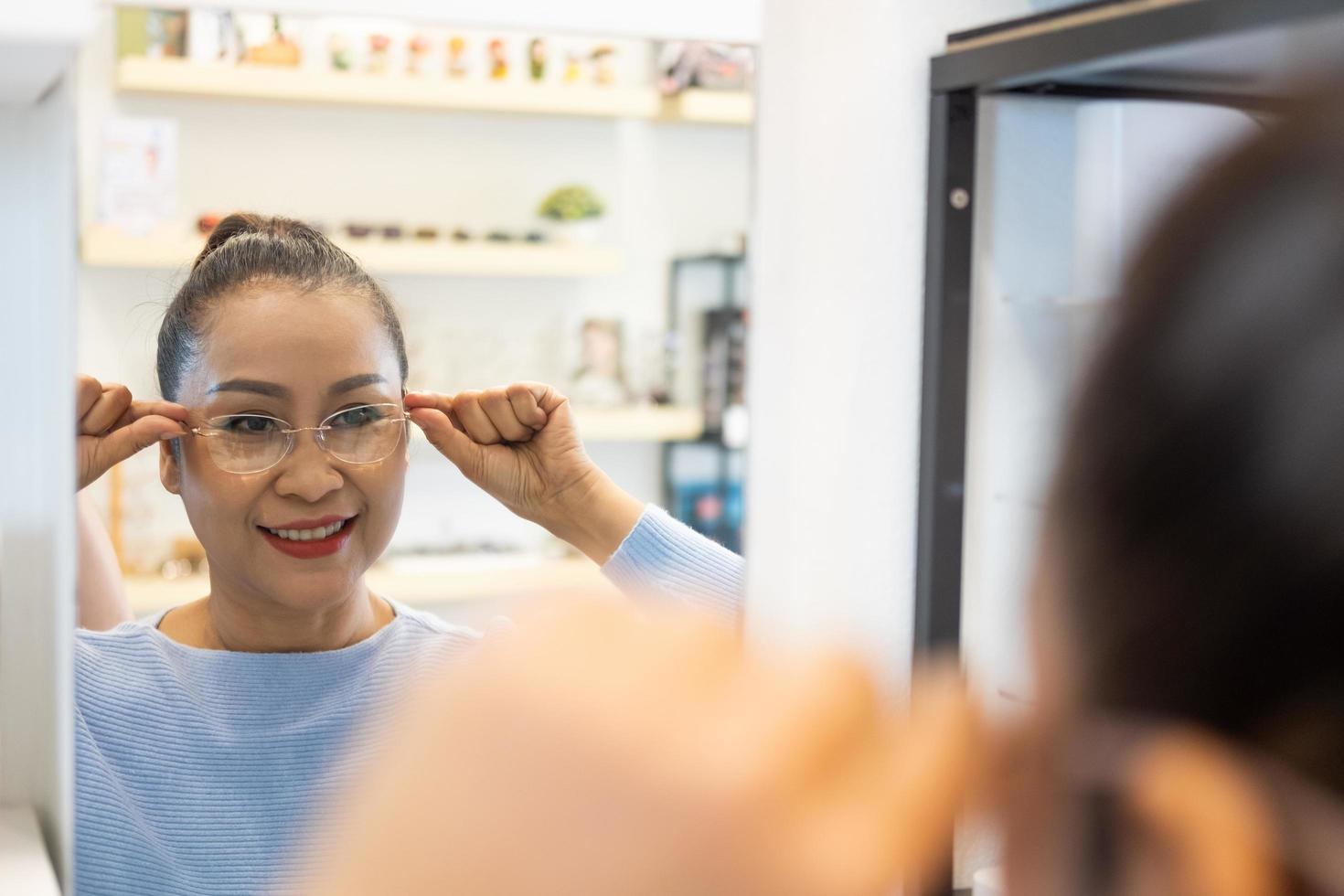 selectief focus Bij de spiegel. senior ouderling Aziatisch Dames glimlach en kijken Bij de spiegel terwijl kiezen mooi bril kader beproeving binnen van optisch winkel, op te slaan. oog zicht voor ouderling, gezondheidszorg. foto