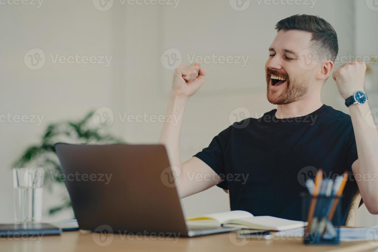 gelukkige jonge freelancer die een winnend gebaar toont terwijl hij thuis op zijn werkplek zit foto