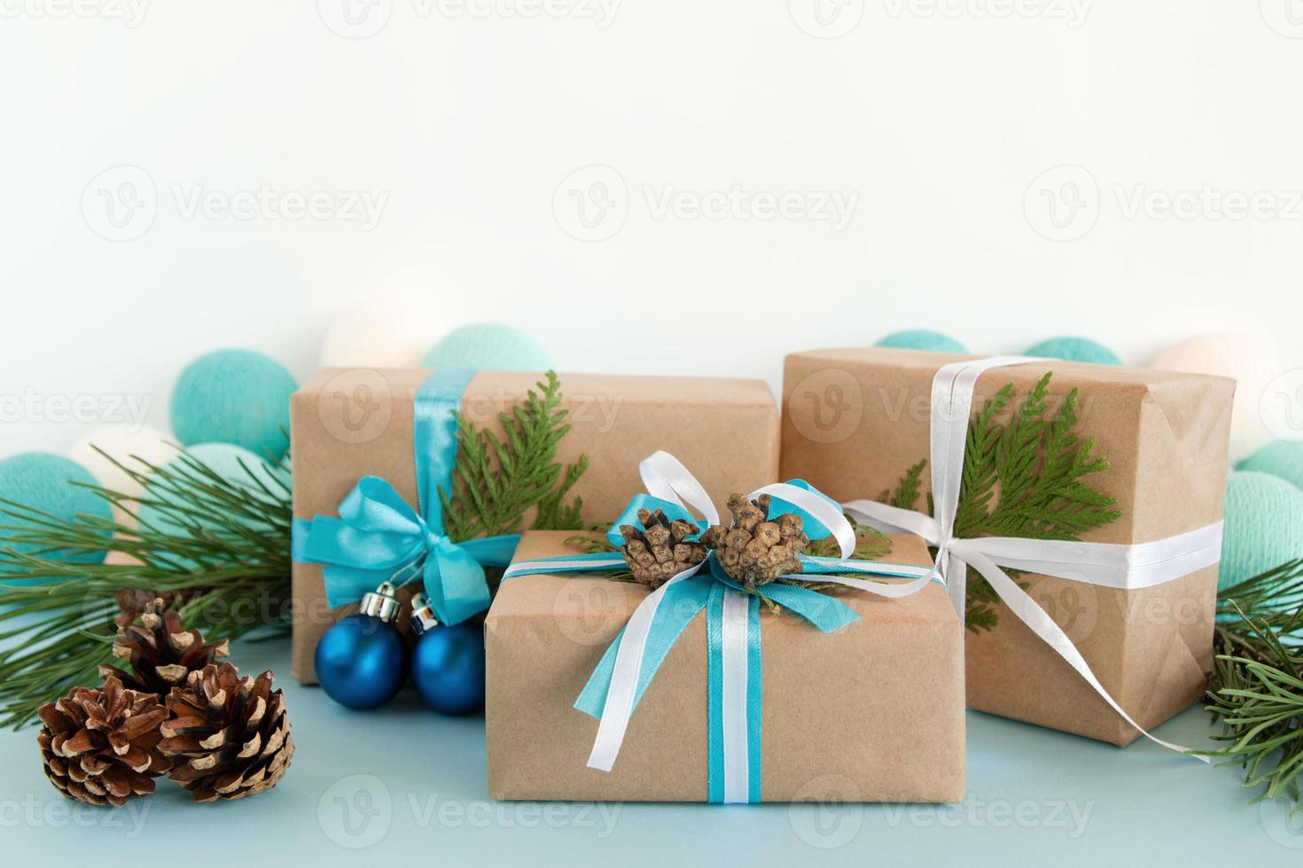 drie Kerstmis geschenk dozen verpakt van ambacht papier, blauw en wit linten, versierd van Spar takken, pijnboom kegels en Kerstmis ballen Aan de blauw en wit achtergrond met Kerstmis lichten. foto