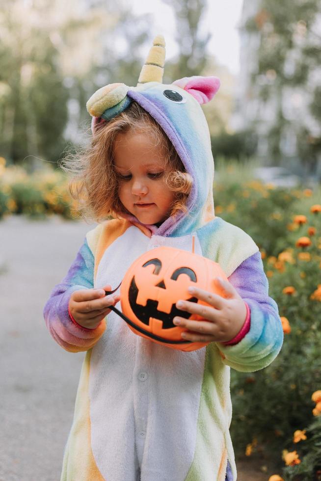 schattig weinig meisje in een regenboog eenhoorn kostuum voor halloween gaat naar verzamelen snoepgoed in een pompoen mand in een woon- Oppervlakte. kind wandelingen in de buitenshuis. truc of traktatie. levensstijl. kigurumi foto
