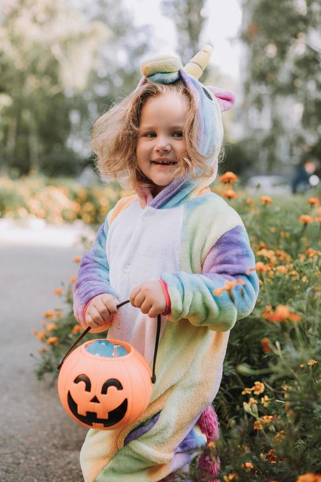 schattig weinig meisje in een regenboog eenhoorn kostuum voor halloween gaat naar verzamelen snoepgoed in een pompoen mand in een woon- Oppervlakte. kind wandelingen in de buitenshuis. truc of traktatie. levensstijl. kigurumi foto