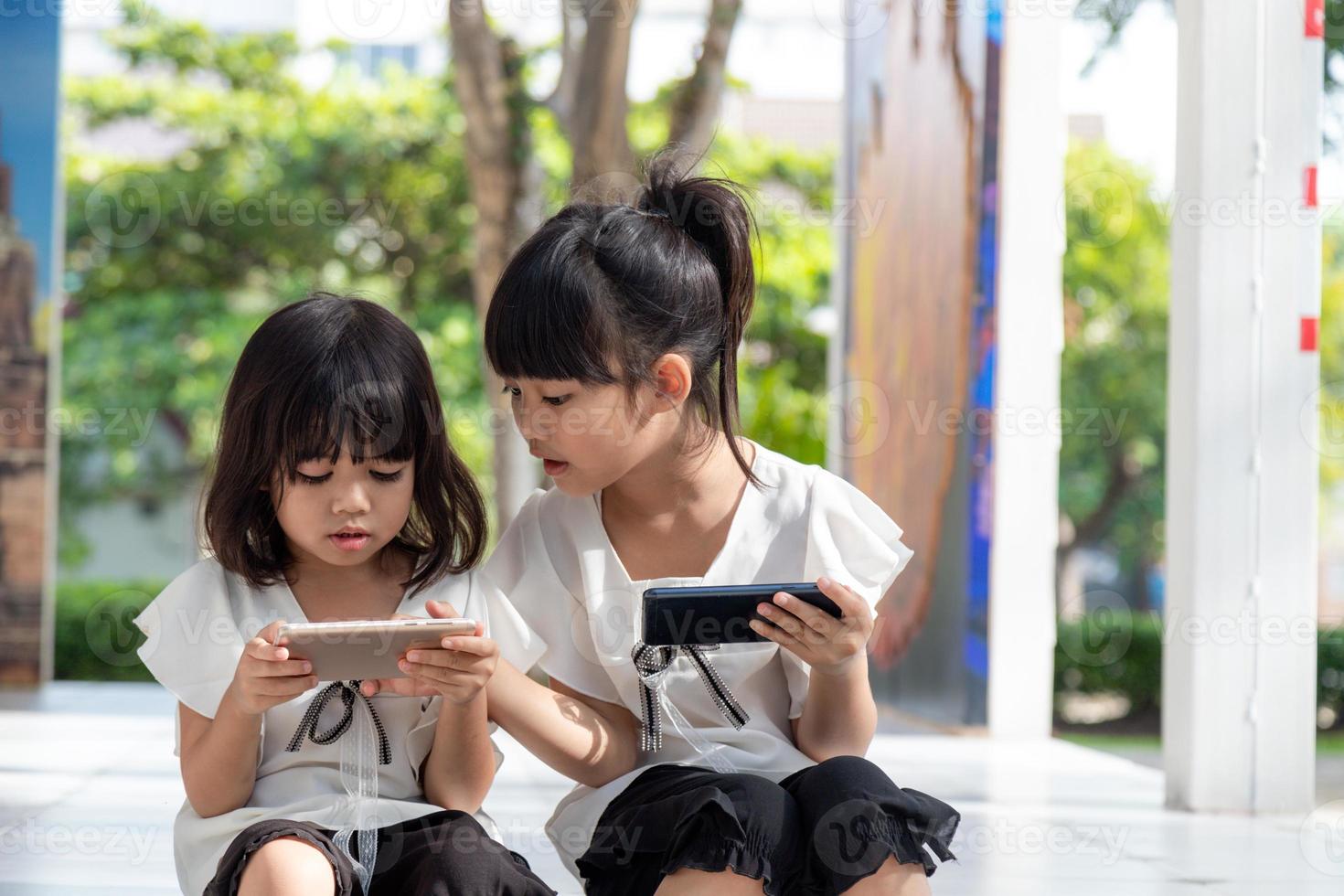 concept kinderen en gadgets. twee weinig meisjes broers en zussen zussen kijken Bij de telefoon en glimlach. ze houden een smartphone kijk maar videos, leren, Speel spellen, spreken online. internet voor kinderen. ouderlijk controle foto