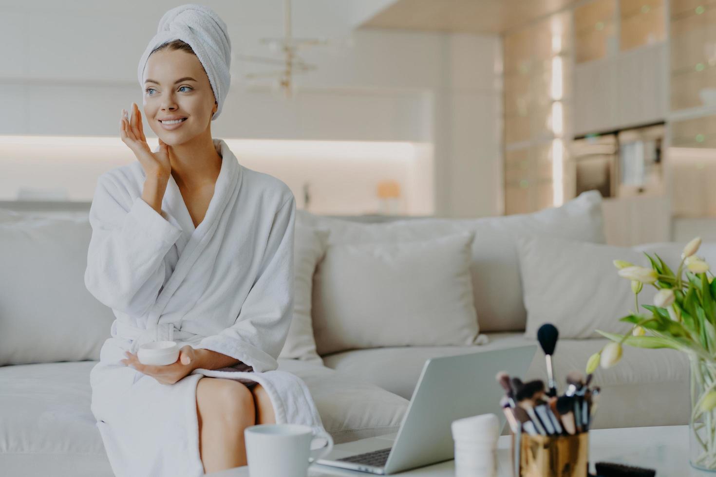 mensen schoonheid en huidverzorging concept. tevreden vrouw brengt gezichtscrème aan geniet van gezichtsbehandelingen kijkt opzij glimlacht zachtjes gebruikt cosmetisch product poseert op de bank voor een geopende laptop foto