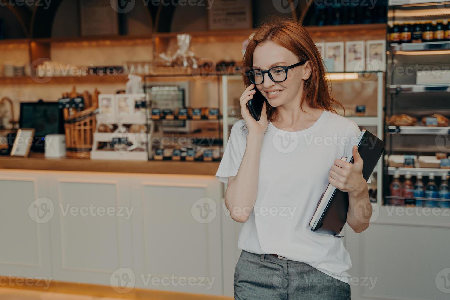 jonge, aangename roodharige vrouw freelancer die aan de telefoon praat en glimlacht terwijl ze café-koffieshop verlaat foto